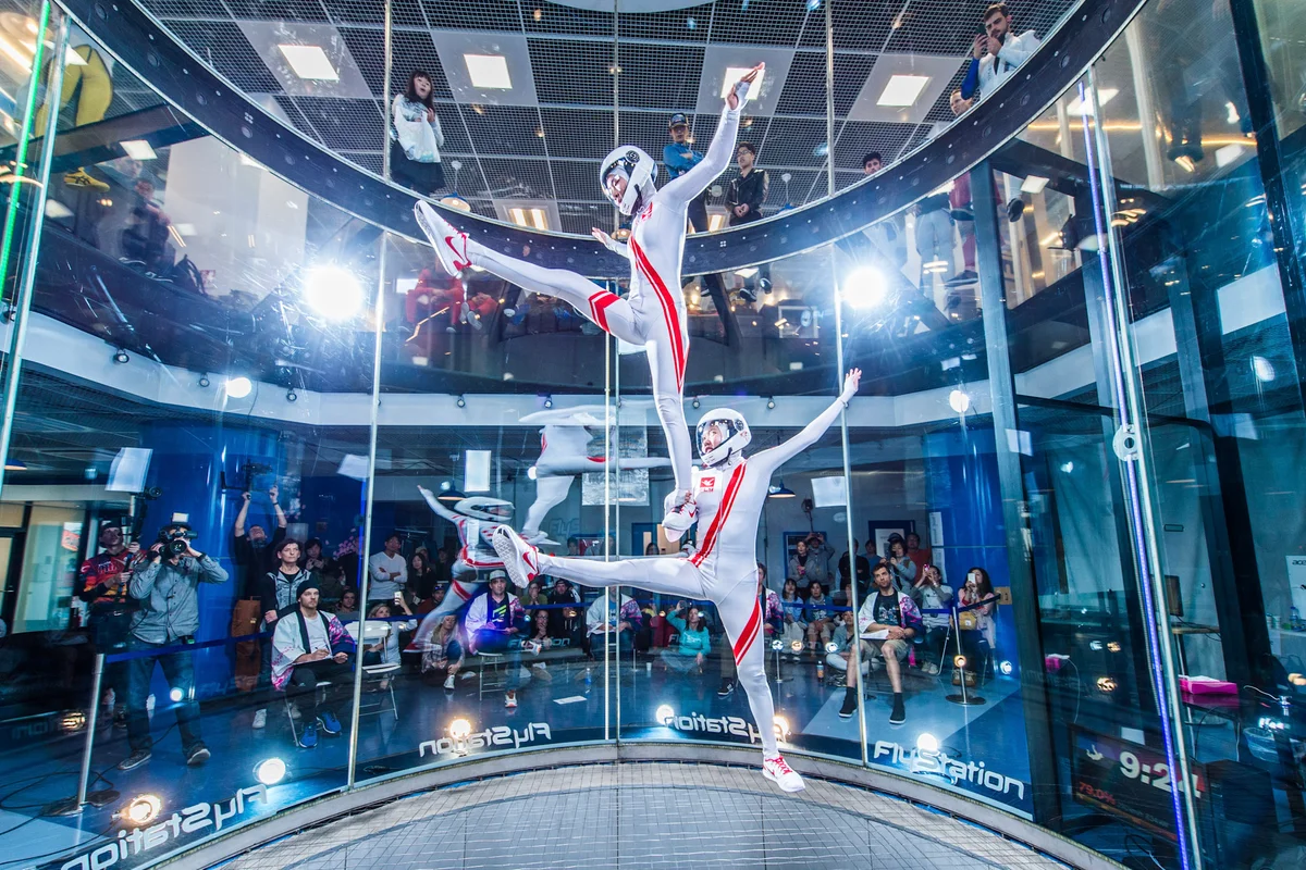 İki uçucu, FlyStation Japan'daki TT45 PRO uçuş odası içinde senkronize bir freestyle manevrası gerçekleştiriyor. Sporcular, düşük sürtünmeli çelik kablolu güvenlik ağı üzerinde dengeli bir formasyon sergiliyor. 4,5 metre çapındaki rüzgâr tüneli, çerçevesiz çok katmanlı cam (multilayer glass) duvarlara sahiptir ve izleyicilerin performansı hem zemin kattan hem de üst asma kattan izlemesine olanak tanır. 2017 yılında devreye alınan (commissioned) bu tesis, gelişmiş dynamic ve artistik uçuş disiplinleri için uygun, kararlı ve yüksek hızlı hava debisi (airflow) ile Sakura Cup gibi profesyonel yarışmaları desteklemektedir.