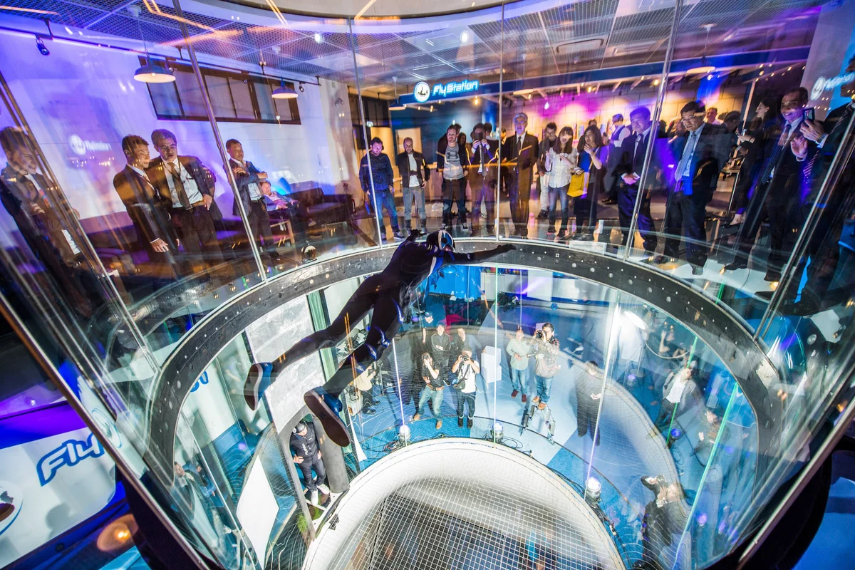 O voador profissional Leo Volkov realiza manobras dentro da câmara de voo TT45 PRO na FlyStation Japan. A perspectiva de cima para baixo revela as paredes cilíndricas de vidro sem moldura e a rede de segurança de aço inoxidável na base da coluna de fluxo de ar. Os espectadores observam o voo a partir do piso de vidro estrutural que circunda a seção superior da câmara. Este túnel de vento de 4,5 metros de diâmetro apresenta o design exclusivo de vidro multicamadas redondo da TunnelTech, garantindo visibilidade e segurança para manobras em alta velocidade.