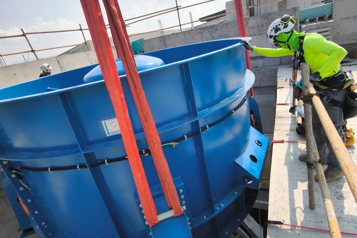 Bauarbeiter leiten die Installation einer Hochleistungs-Axialventilatoreinheit für einen TunnelTech TT43 Smart Windkanal beim Projekt China Flight Town. Das blaue Ventilatorgehäuse aus Stahl, aufgehängt an Schwerlast-Krangurten, wird in den vertikalen Betonschacht des Rückführkanals abgelassen. Unterhalb des Gehäuses ist das Laufrad mit CFK-Schaufeln sichtbar, das für einen effizienten Betrieb im geschlossenen Luftkreislauf ausgelegt ist.