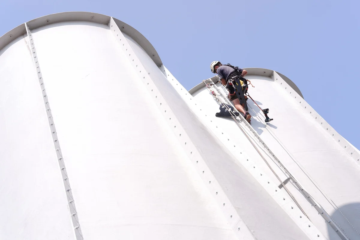 Isang installation technician ng TunnelTech ang nagsasagawa ng assembly work sa labas ng mga seksyon ng diffuser ng patayong wind tunnel gamit ang rope access equipment. Ang malalaking cylindrical duct segments ay nagdudugtong sa pamamagitan ng vertical bolted flanges upang mabuo ang itaas na bahagi ng aerodynamic loop. Ang teknik na ito ng assembly ay nagbibigay-daan sa team na ma-secure ang mga dugtungan at ma-inspeksyon ang istruktura sa matataas na patayong bahagi nang hindi gumagamit ng mabibigat na scaffolding.