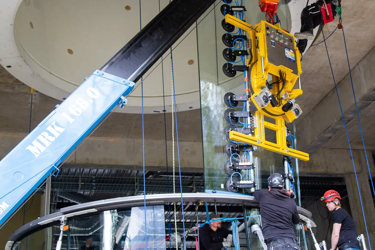 Una grúa araña especializada equipada con un elevador por vacío de alta resistencia posiciona un gran panel de vidrio curvo para la cámara de vuelo TT45 PRO. Escaladores industriales suspendidos del anillo de hormigón superior guían el vidrio multicapa fonoabsorbente hacia la estructura de acero, mientras que los técnicos en los andamios alinean la base. Este proceso de montaje en las instalaciones de Wind Alps en Francia construye la zona de vuelo cilíndrica y transparente del túnel de circuito cerrado.