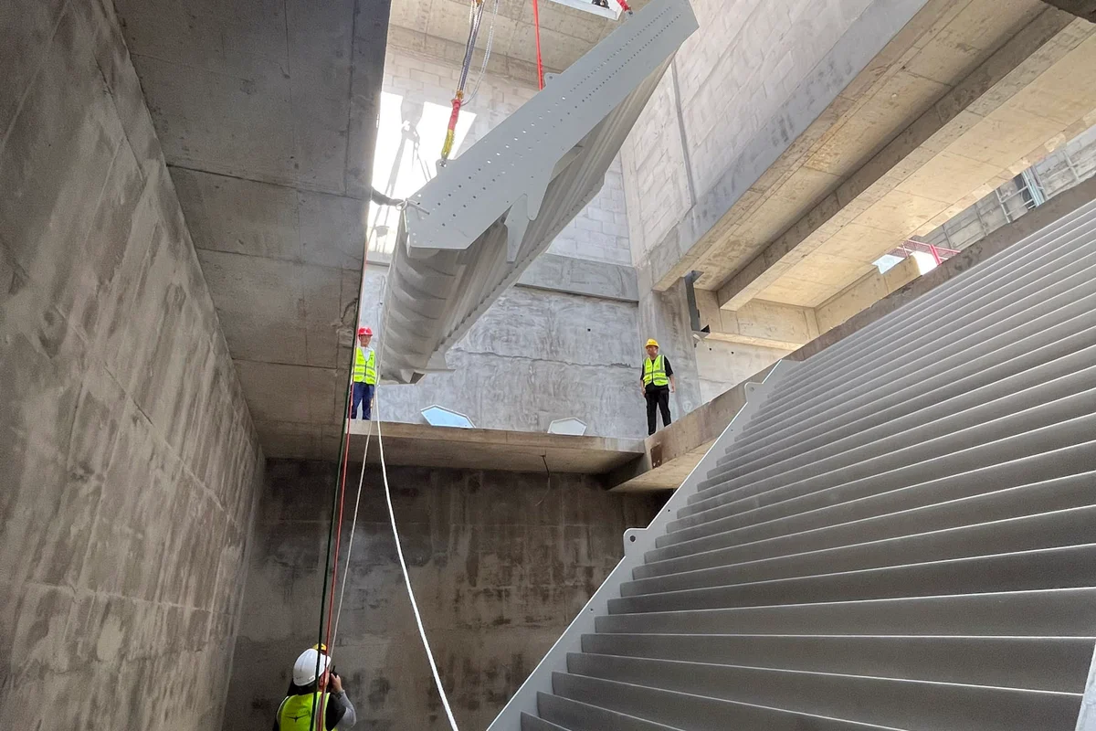 Técnicos orientam a instalação de aletas guia com arrefecimento integrado na estrutura de betão da conduta do túnel de vento TT43 Smart na China Flight Town. Uma grua baixa o segmento de aleta de alumínio através de uma abertura superior para a conduta de retorno. Estas aletas guia incorporam canais internos de refrigerante para funcionar como permutadores de calor, gerindo a temperatura do ar enquanto redirecionam o fluxo de ar em 90 graus com mínima turbulência e perda de pressão.