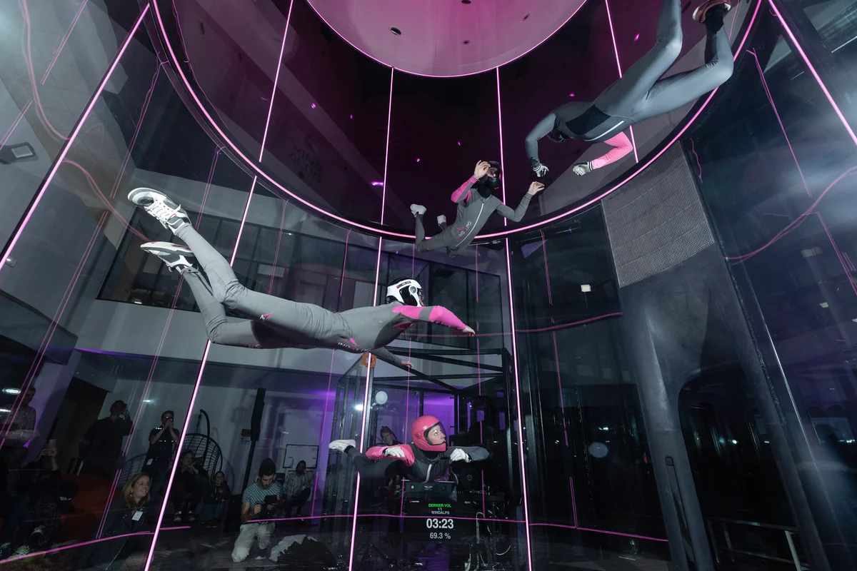 High-performance team training in action: four flyers share the air inside the TT45 PRO flight chamber at Wind Alps, France. This 8-meter tall section features perfectly round, multilayer noise-absorbing glass with vertical lighting, ensuring a quiet environment for spectators while offering a clear view of the action. A telemetry display directly on the glass surface provides real-time data on flight duration and wind speed settings.