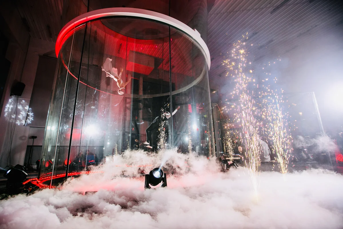 Esta imagem espetacular da Kuzbass Arena mostra a versatilidade da câmara de voo TT52 Pro. Enquanto fogos de artifício e neblina criam uma atmosfera dramática fora do vidro, uma flyer de vestido branco executa uma manobra precisa de head-down no interior, acompanhada por um segundo atleta. Isso demonstra como a tecnologia TunnelTech suporta entretenimento de alto nível e eventos artísticos ao lado do esporte profissional.