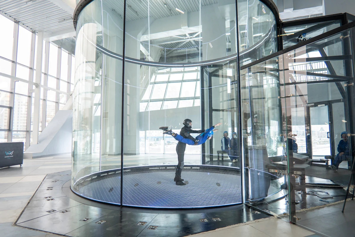 Professional instruction in action inside the TT52 Pro wind tunnel at Kuzbass Arena. This massive 5.2-meter flight chamber sets the standard with its frameless, noise-absorbing glass that ensures perfect visibility and uniform airflow. You can see the instructor spotting a belly flyer above the low-drag steel cable safety net, with the seamless glass airlock entry visible to the right.