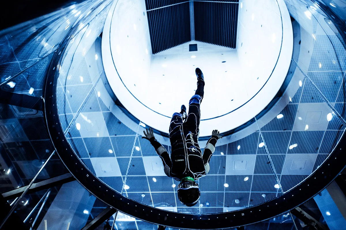 En professionell flygare utför en head-down manöver inuti glasflygkammaren i TT45 PRO vindtunneln på FlyStation Japan. Perspektivet underifrån avslöjar övergången från den transparenta flygsektionen till diffusorn och det övre kanalhörnet utrustat med parallella ledskovlar. Flygaren bär en dräkt med logotyp medan han navigerar i det snabba vertikala luftflödet. Kammaren på 4,5 meter i diameter har böjda paneler av flerskiktsglas utformade för synlighet och säkerhet under avancerade flygoperationer.