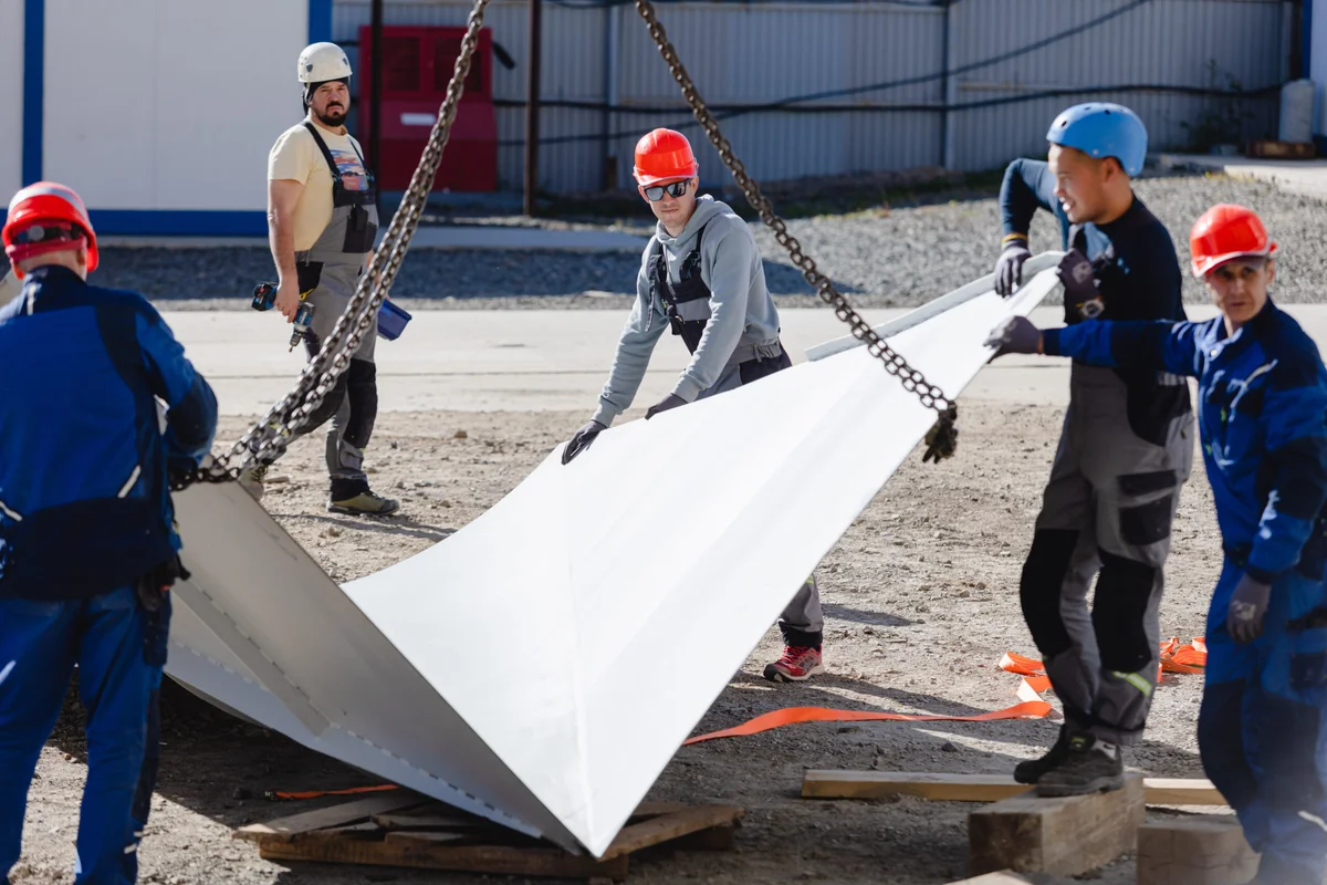 Uma equipa de instalação especializada da TunnelTech manobra um grande painel de difusor branco suspenso por correntes de grua. A equipa guia a secção angular de conduta compósita de FRP para a posição de montagem no estaleiro de obra. Este componente estrutural forma uma parte crítica do circuito aerodinâmico do túnel de vento, especificamente concebido para expandir o caudal de ar e recuperar a pressão a jusante da câmara de voo. Os trabalhadores usam equipamento de proteção individual padrão enquanto manuseiam a carga pesada.