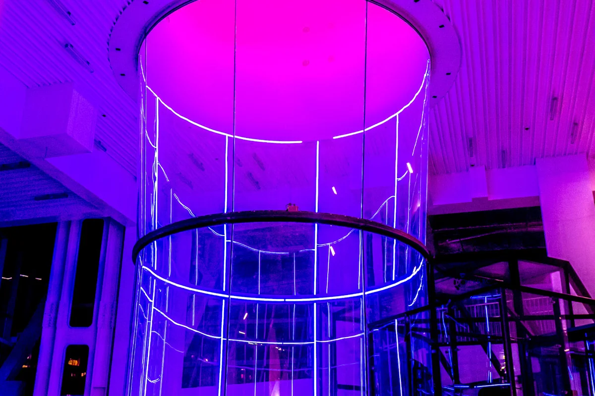 Vue imprenable sur le TT52 Pro de la Kuzbass Arena, mettant en valeur son imposante chambre de vol de 5,2 mètres de diamètre. La section cylindrique, construite en verre multicouche insonorisant et baignée d'une lumière violette, se connecte parfaitement au conduit de retour supérieur. Sur la droite, on distingue l'antichambre vitrée et le système de sas d'accès. Cette installation repose sur une conception à recirculation à boucle unique, optimisée pour un flux important de volants et l'entraînement professionnel.