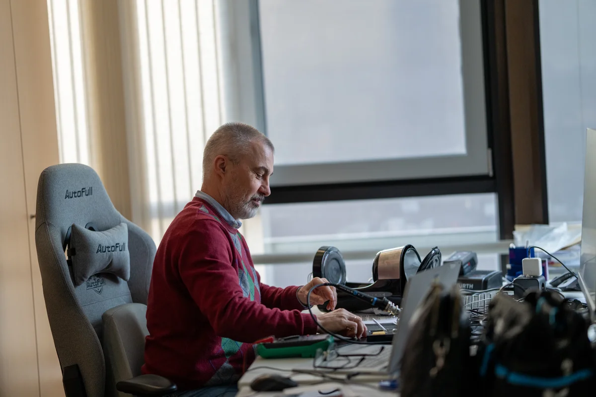 TunnelTech inženieris strādā pie galda, kas aprīkots elektronikas montāžai un testēšanai. Darba vietā ir lodāmurs, digitālais multimetrs un precīzijas instrumenti, ko izmanto aparatūras apkopei vai izstrādei. Inženieris koncentrējas uz shēmas komponentiem, atbalstot uzņēmuma iekšējo vadības elektronikas un automatizācijas sistēmu ražošanu vēja tuneļa operācijām.