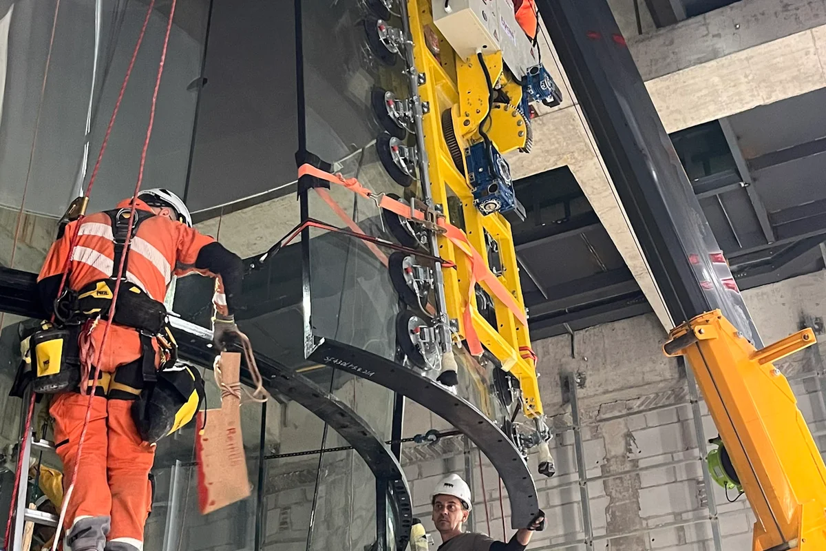 Tekniker installerar en böjd glaspanel för flygkammaren i en TT43 Smart vindtunnel vid China Flight Town-projektet. En kraftfull vakuumlyftare fäst vid en kran positionerar den flerskiktiga ljudabsorberande glassektionen i stålramen. Installationsteamet använder repåtkomstteknik och stegar för att rikta in det kemiskt förstärkta glaset, vilket säkerställer exakt passform för flygområdet med en diameter på 4.3 meter.