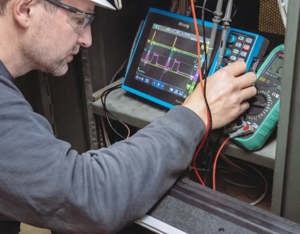 En servicetekniker udfører elektrisk diagnostik på vindtunnelens industrielle automations- og styresystemer. Ingeniøren bruger et Micsig tablet-oscilloskop til at måle signalbølgeformer og verificere de elektroniske komponenters ydeevne. Det åbne udstyrsrack indeholder DIN-skinne monterede interfacemoduler og ledningsterminaler. Denne vedligeholdelsesaktivitet understøtter pålideligheden af den interne kontrolelektronik og software udviklet af TunnelTechs ingeniørteam.