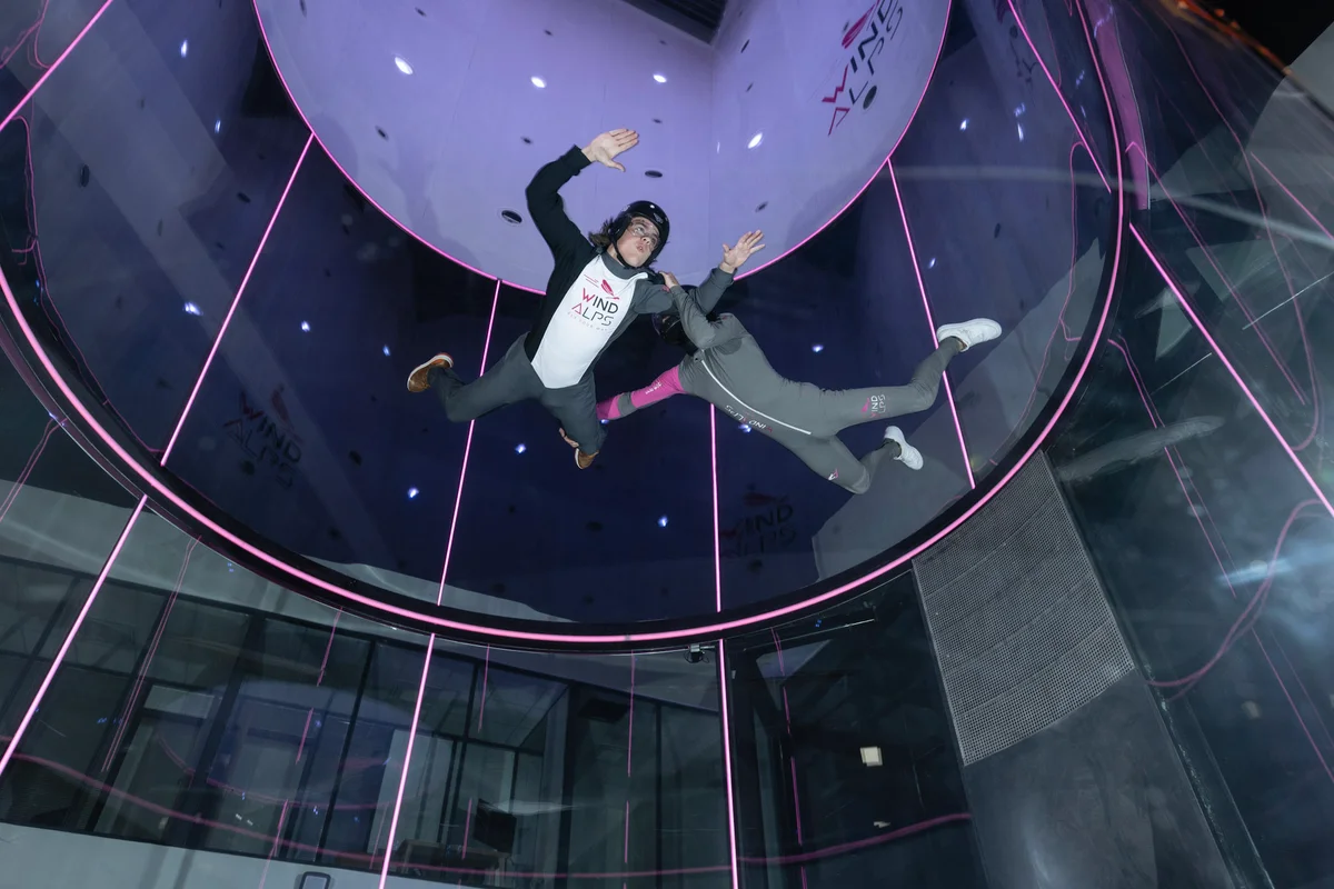 Inside the TT45 PRO wind tunnel at WindAlps, France. An instructor guides a beginner through the taxi-flight maneuver. The signature conical glass section maximizes visible flying space, while the high-tension safety net ensures a secure training environment.