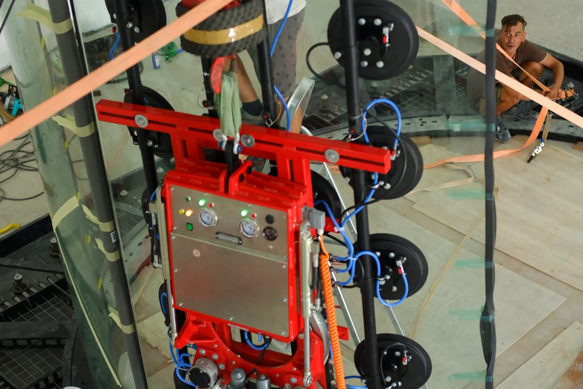 A equipa técnica está a instalar um painel de vidro multicamadas curvo para o túnel de vento TT45 PRO nas instalações da Brimob, na Indonésia. Um elevador a vácuo de alta resistência segura a secção transparente enquanto os trabalhadores a guiam para a posição acima da rede de segurança. Assim se constrói a câmara de voo de 4,5 metros de diâmetro, utilizando vidro quimicamente reforçado para garantir elevada segurança contra impactos e clareza ótica em ambientes de treino profissional.