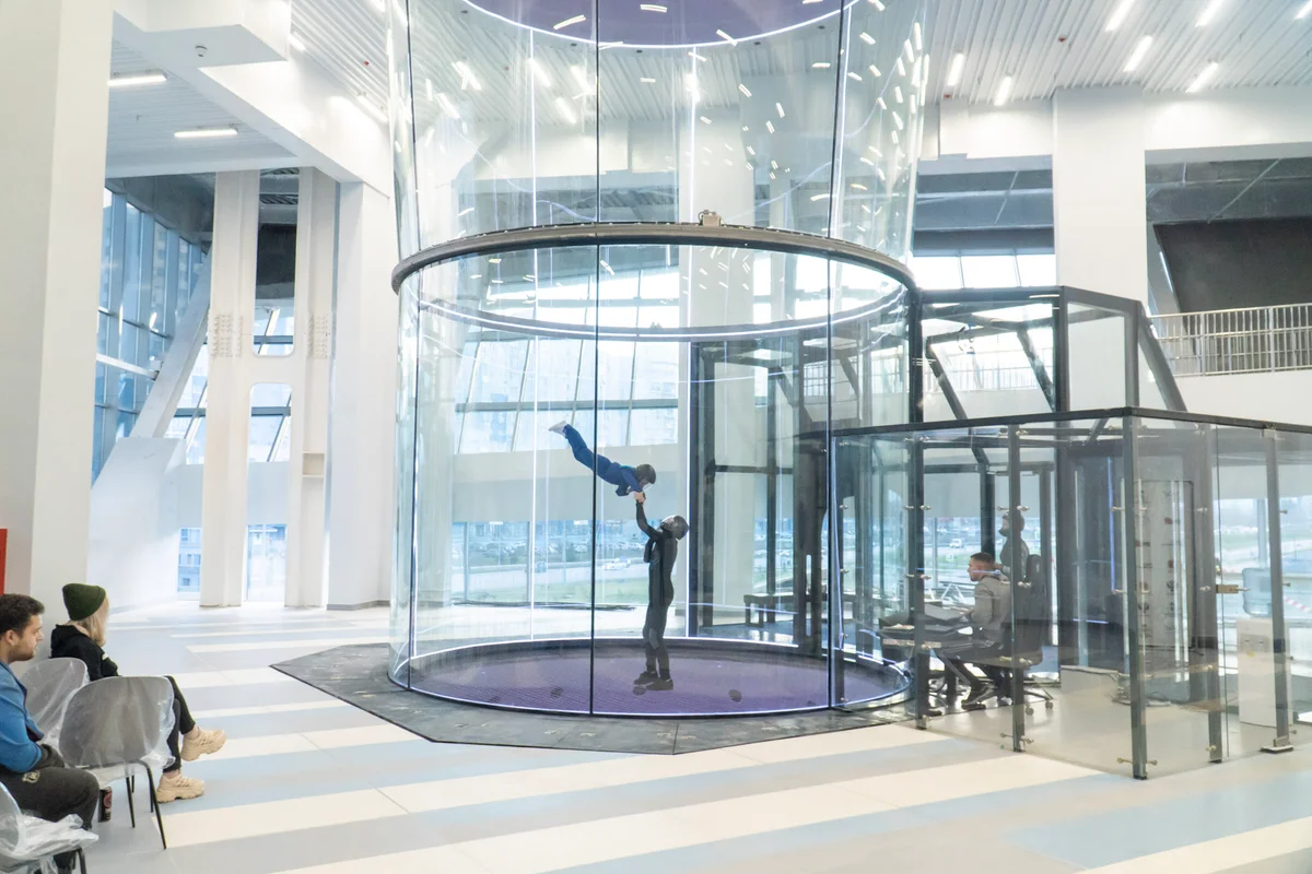 Seorang instruktor membantu pelayang muda dalam posisi belly flying di dalam ruang penerbangan (flight chamber) TT52 Pro di Kuzbas Arena. Instalasi berdiameter 5.2 meter ini dilengkapi dinding kaca berlapis (multilayer glass) tanpa bingkai untuk keselamatan dan visibiliti maksimum. Di sebelah kanan, operator mengawal aliran udara (airflow) dari stesen kerja yang terletak bersebelahan dengan airlock kaca.