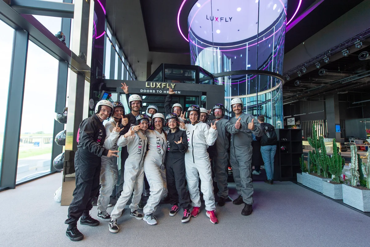 A group of flyers wearing flight suits and helmets stands in the interior of the Luxfly wind tunnel facility in Luxembourg, preparing for a flight session. The participants pose in front of the glass flight chamber entrance, labeled "Luxfly Doors to Heaven," within the modern facility.