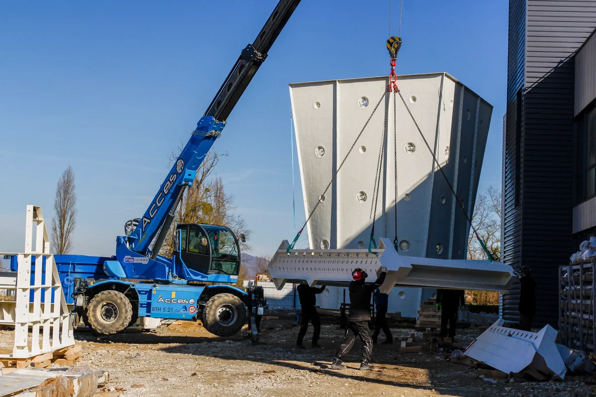 De foto toont een keerschoepenmodule die door een kraan uit het gebouw wordt gehesen. Op de achtergrond is een diffuser van de vliegruimte met openingen voor verlichting zichtbaar. Keerschoepen worden gebruikt om de luchtstroom binnen het recirculatiecircuit om te leiden, waardoor turbulentie en geluid worden verminderd.