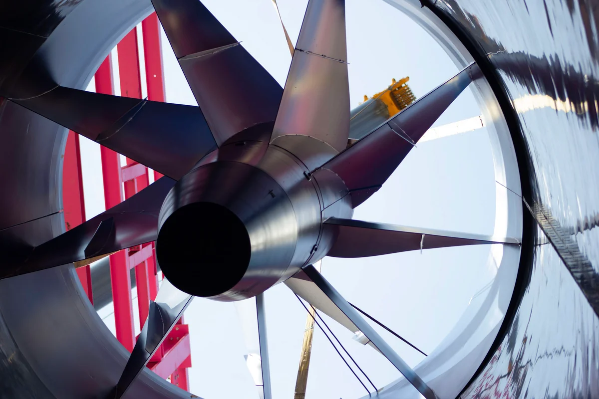 Foto ini mengabadikan pemasangan unit kipas aksial utama untuk terowongan angin Luxfly di Luksemburg. Rakitan ini memiliki rotor berdiameter besar dengan bilah serat karbon yang dipasang pada sumbu vertikal. Sebuah derek memposisikan komponen kipas di dalam saluran udara (ductwork), dikelilingi oleh struktur pendukung baja merah fasilitas tersebut. Komponen ini menggerakkan aliran udara untuk sistem TunnelTech TT45 PRO.