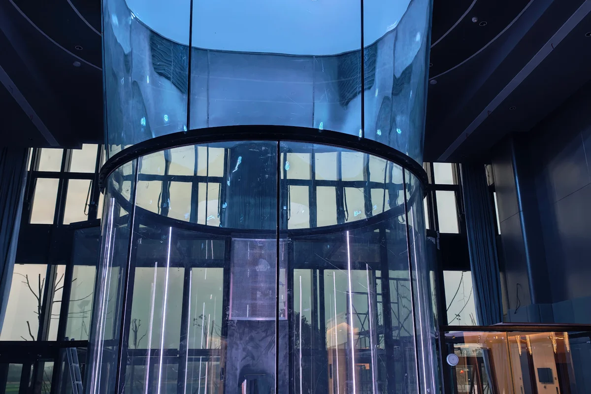 China Flight Town施設のTT43 Smartフライトチャンバーは、直径4.3メートルの円筒形ガラスセクションを特徴としています。完全に丸い多層ガラスパネルが透明な壁を形成し、垂直のLEDストリップによって照らされています。この構造は、飛行空間の上にある上部ディフューザーとFRP複合ダクトに接続しています。底部では、安全ネットが低抵抗のスチールケーブルを使用しています。この設備は、SMARTflyシリーズで利用可能な拡張ガラス構成を採用しており、近代的な商業ビルの内部に設置されています。