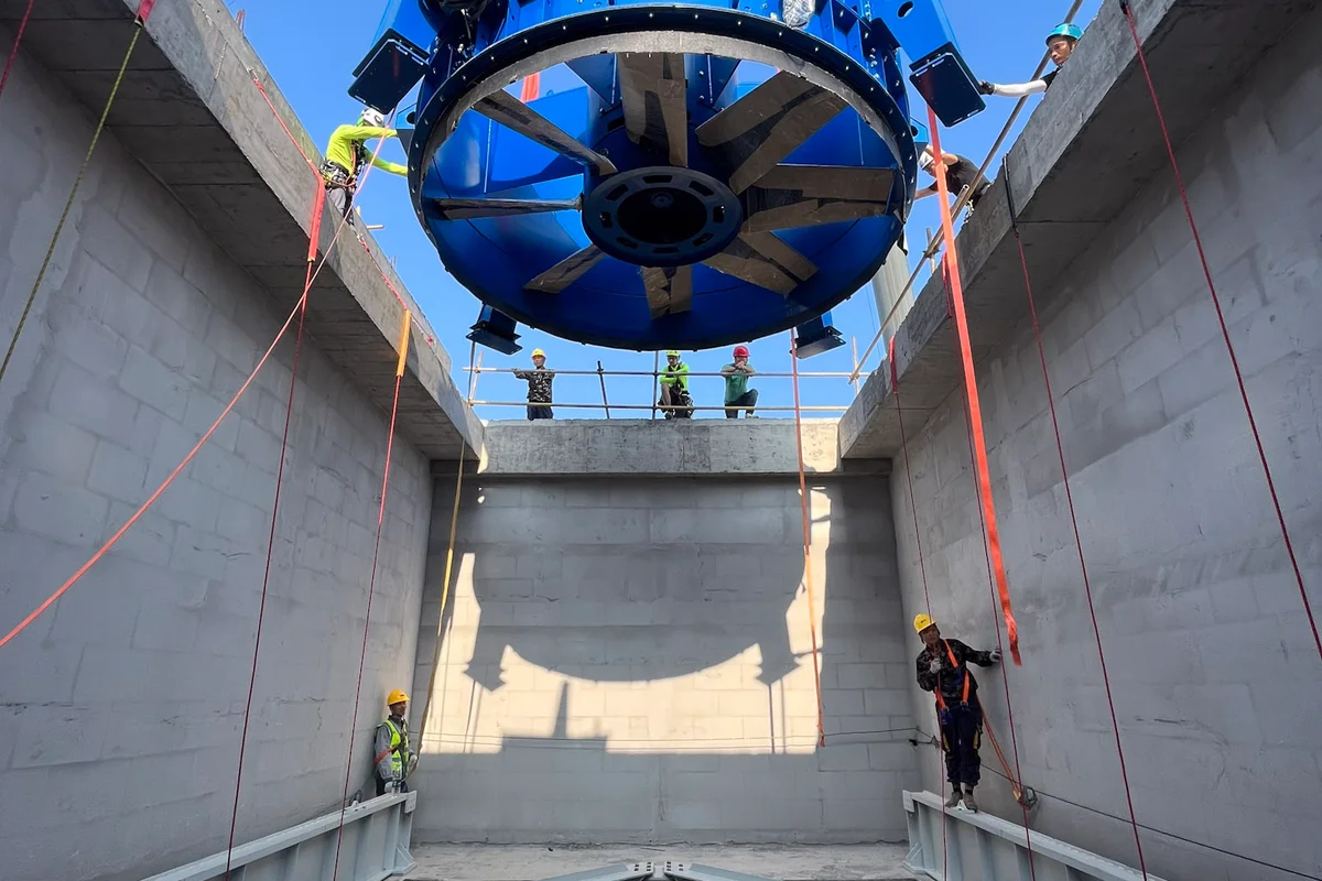 China Flight Town施設にて、大型クレーンが青色のEVG製軸流ファン（Axial fan）ユニットをTT43 Smart風洞（Wind tunnel）の垂直コンクリート製リターンダクト（Return duct）内に吊り下ろしています。カーボンファイバーブレード（Carbon-fiber blades）を備えたファン組立品は、シャフトの底部に配置された固定式の案内羽根（Guide vanes）構造に向かって下降しています。据付技術者がロープアクセス技術を使用して負荷を誘導し、コンクリート筐体内での正確な位置合わせを確実にしています。この据付工程により、主要な気流（Airflow）生成コンポーネントがリターンループ構造内に配置されます。