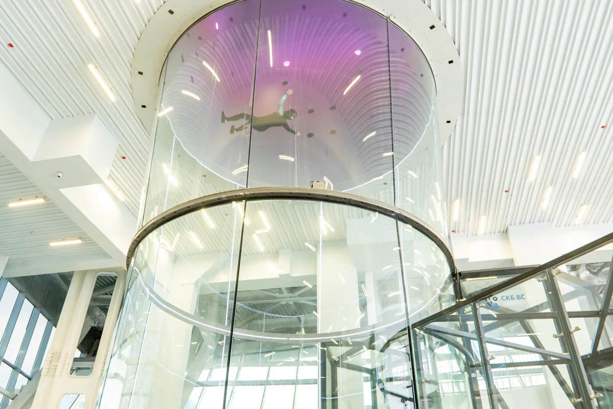 The TT52 Pro wind tunnel at Kuzbass Arena features a 5.2-meter diameter flight chamber constructed from perfectly round, multilayer noise-absorbing glass. You can see a flyer navigating the airflow while the operator manages system controls from the external workstation. This facility utilizes a single-loop design optimized for professional training, providing unobstructed visibility through the frameless glass sections.
