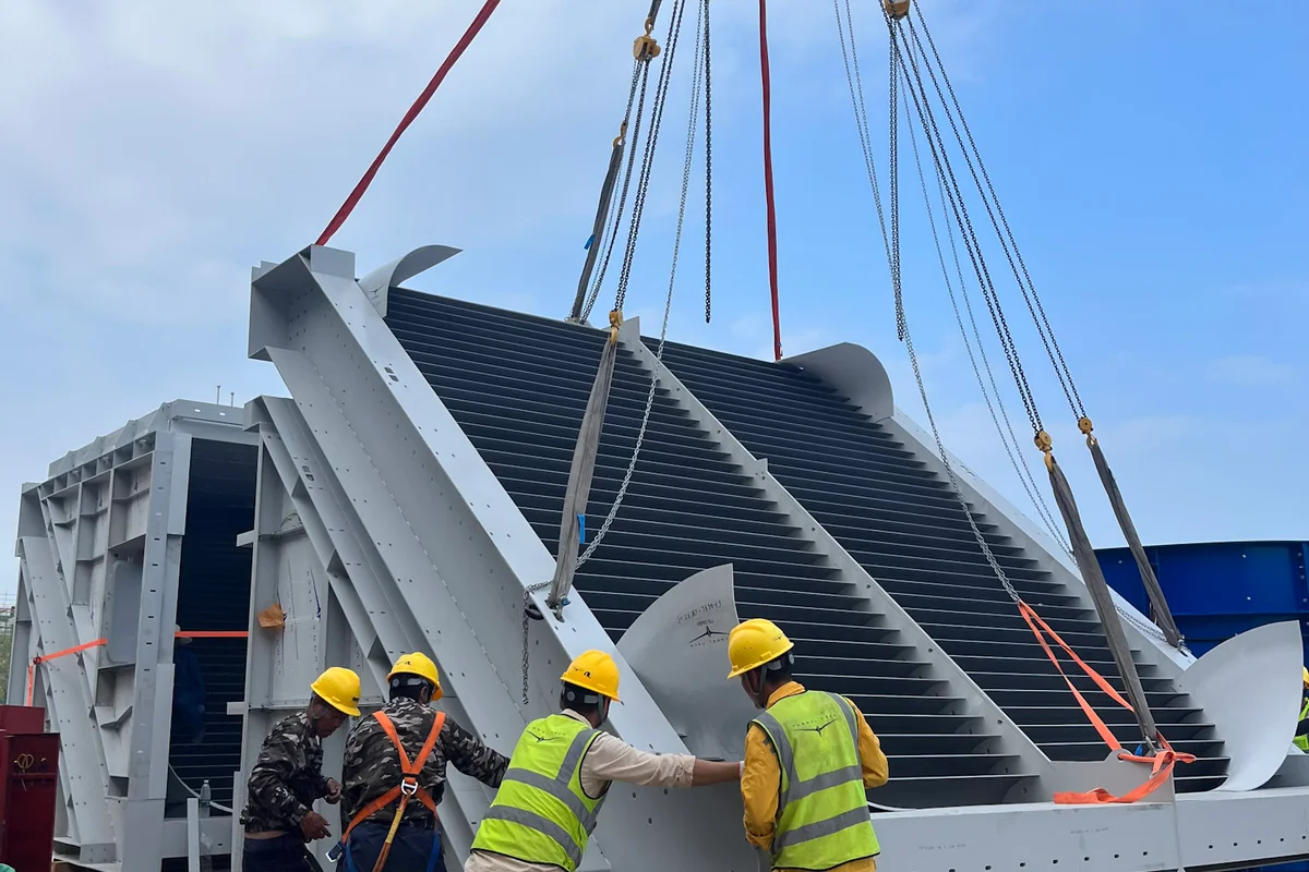 Một chiếc cần cẩu đang nâng một đoạn cánh hướng dòng trong quá trình lắp đặt hầm gió TT43 Smart tại dự án China Flight Town. Nhân viên xây dựng đang dẫn hướng khối thiết bị khung thép, chứa các kênh dẫn chất làm mát tích hợp cho hệ thống làm mát chủ động của cơ sở. Các cánh này hoạt động như bộ trao đổi nhiệt để điều chỉnh nhiệt độ luồng khí trong khi đổi hướng gió qua góc của vòng ống dẫn tuần hoàn.