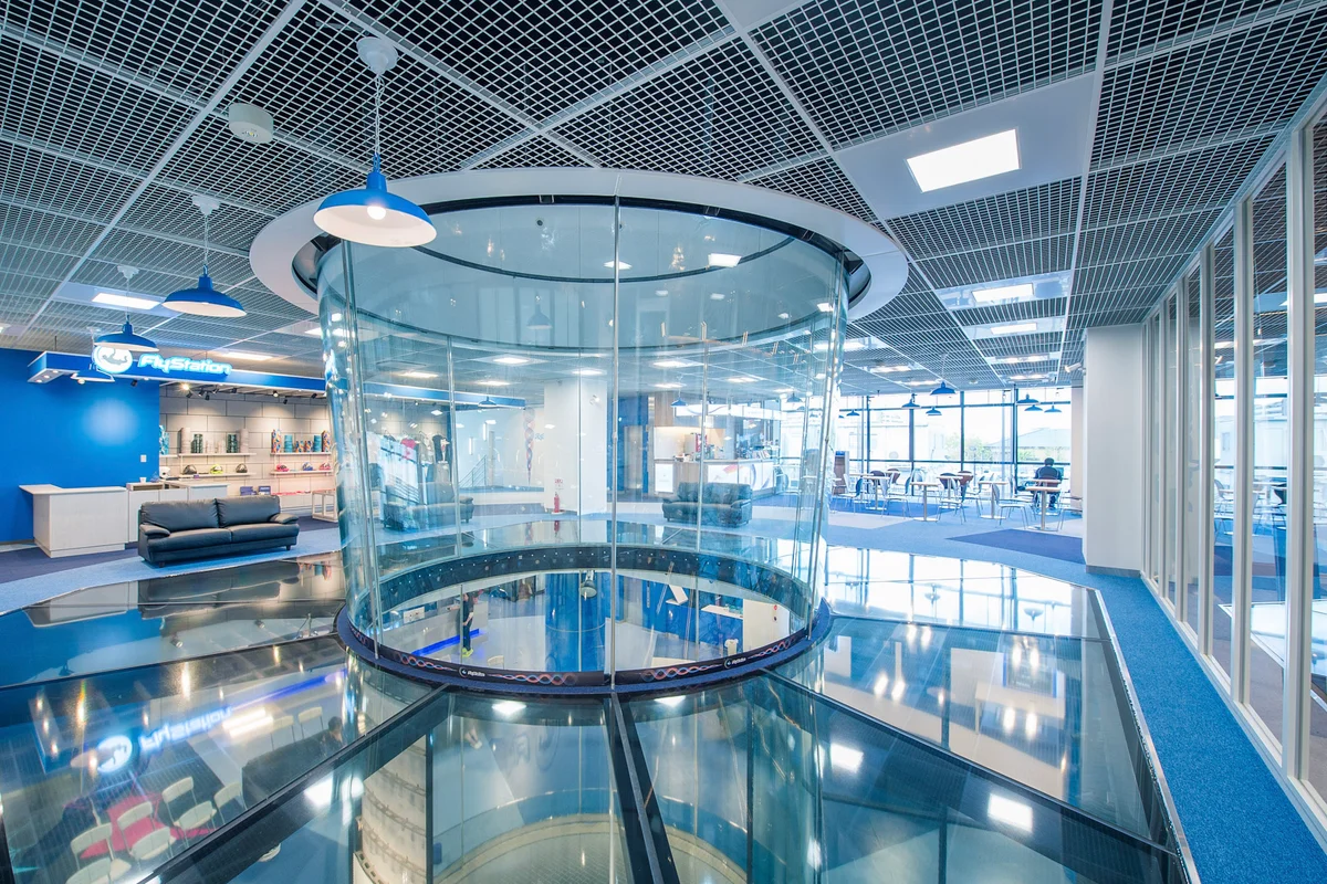 Denne indvendige visning af FlyStation Japan viser den øvre sektion af et TunnelTech TT45 Pro flyvekammer. Et strukturelt glasgulv omkranser tunnelen og giver vertikalt udsyn til flyveområdet og de nedre niveauer. Installationen anvender buede flerlag glas-paneler til flyvekammerets vægge. Det omkringliggende kommercielle område inkluderer en reception med FlyStation-branding, butikshylder og tilskuerpladser, hvilket viser integrationen af vindtunnel-infrastrukturen i anlæggets offentlige zone.
