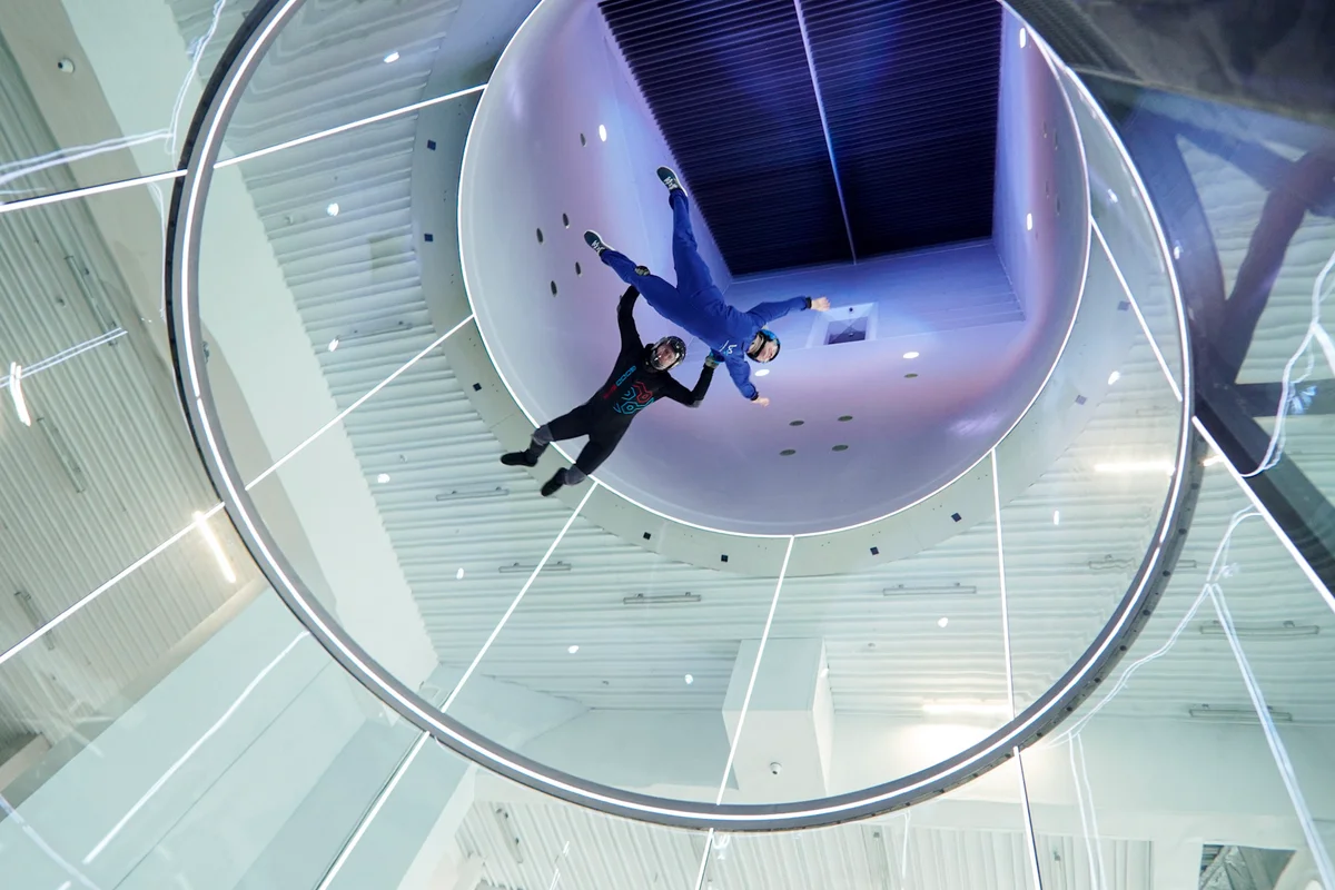 Een verticaal perspectief omhoog door de 5,2 meter brede vliegruimte, waar een instructeur een beginner meeneemt voor een hoge vlucht. De kenmerkende ronde glazen wanden van TunnelTech gaan over in de witte diffuser, met zicht op de keerschoepen bovenin het circuit. Geïntegreerde lichtringen verlichten de ruimte tijdens de gecontroleerde opstijging.