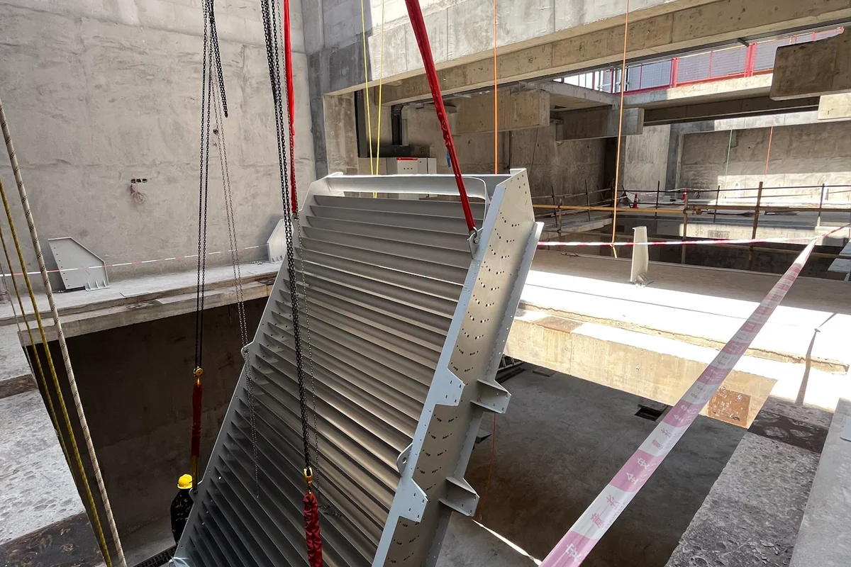 Installation einer Baugruppe aus Umlenkleitblechen mit integrierter aktiver Kühlung beim Projekt China Flight Town. Diese Komponente gehört zu einem TT43 Smart Windkanal und kombiniert Luftstromumlenkung mit Wärmemanagementfunktionen. Die Aluminiumschaufeln enthalten interne Kühlkanäle, um die während des Betriebs erzeugte Wärme abzuführen. Die Baugruppe hängt am Krangeschirr innerhalb der Betonstruktur der Anlage, bereit für die Platzierung in der Ecke des Rückführkanals.