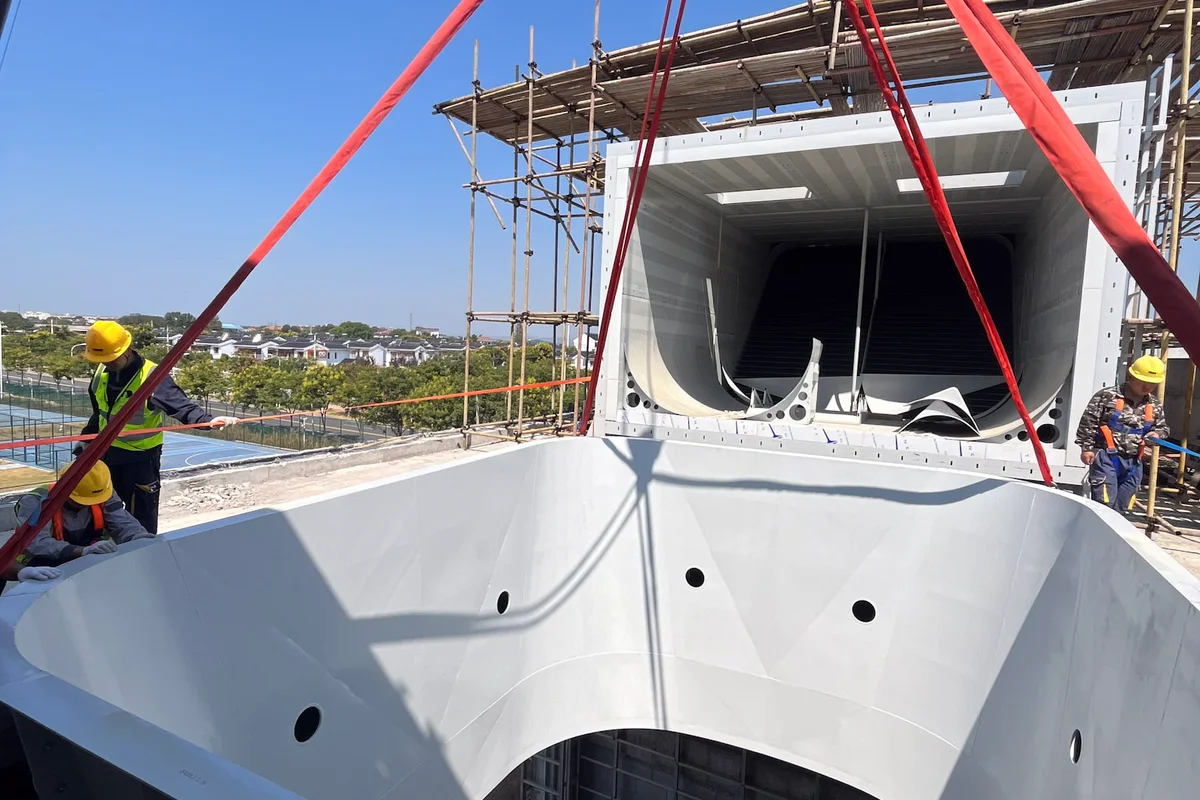 L'installation de la soufflerie TT43 Smart à China Flight Town se poursuit avec le levage d'une section de diffuseur de la chambre de vol. Le composant blanc présente une ouverture circulaire qui se connecte à la zone de vol. En arrière-plan, une section horizontale du conduit de retour équipée d'aubes directrices noires repose sur l'échafaudage en acier. Le personnel de construction gère les sangles de levage pour aligner le lourd segment aérodynamique.