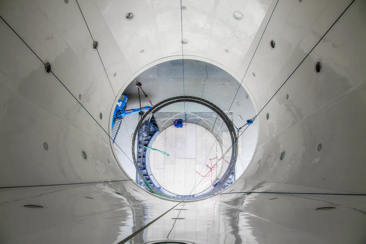 Una prospettiva verticale che guarda verso il basso attraverso la sezione superiore del diffusore di una galleria del vento TT45 PRO presso la struttura Wind Alps. Le pareti a pannelli bianchi del circuito del flusso d'aria presentano porte di accesso circolari e conducono al livello della camera di volo. In basso, catene di manovra e macchinari edili blu assistono nell'installazione dei componenti della camera di volo in vetro e delle flange di collegamento in acciaio.
