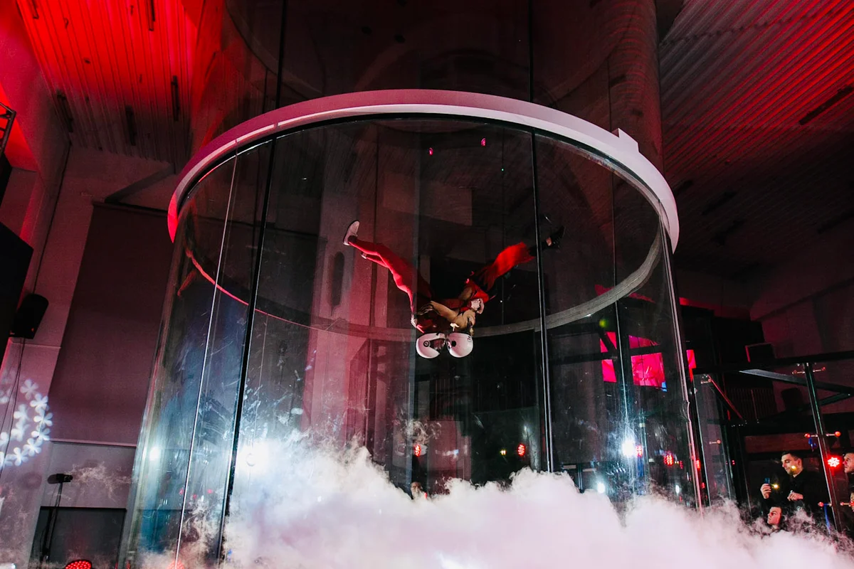 Capturing the art of bodyflight, this pair performs a perfectly synchronized head-down routine inside Kuzbas Arena's TT52 Pro. Hovering above a layer of theatrical smoke, they execute a mid-air kiss while maintaining rock-solid stability in the high-speed airflow. The shot highlights the tunnel's 5.2m frameless glass chamber—a TunnelTech signature—which provides spectators with a crystal-clear view of the action under dramatic red lighting.
