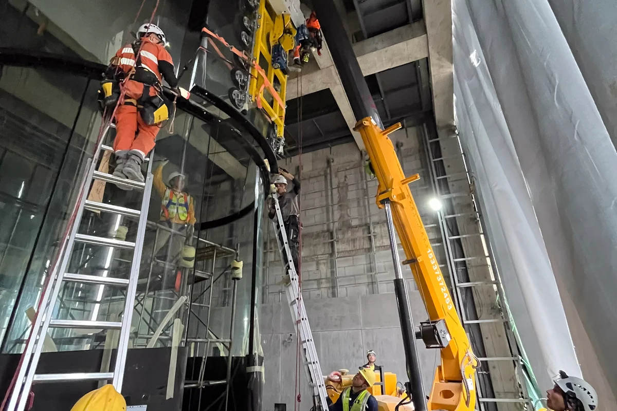 Les techniciens de TunnelTech installent des panneaux de verre multicouche incurvés pour une chambre de vol de soufflerie TT43 Smart sur le site de China Flight Town. Une grue araignée équipée d'un palonnier à ventouses positionne le lourd segment de verre tandis qu'une équipe utilise des techniques d'accès sur cordes et des échelles pour un alignement précis. Le processus d'installation assemble la chambre de vol cylindrique de 4,3 mètres de diamètre, utilisant du verre chimiquement renforcé pour assurer la sécurité structurelle et une aérodynamique de débit d'air constante.