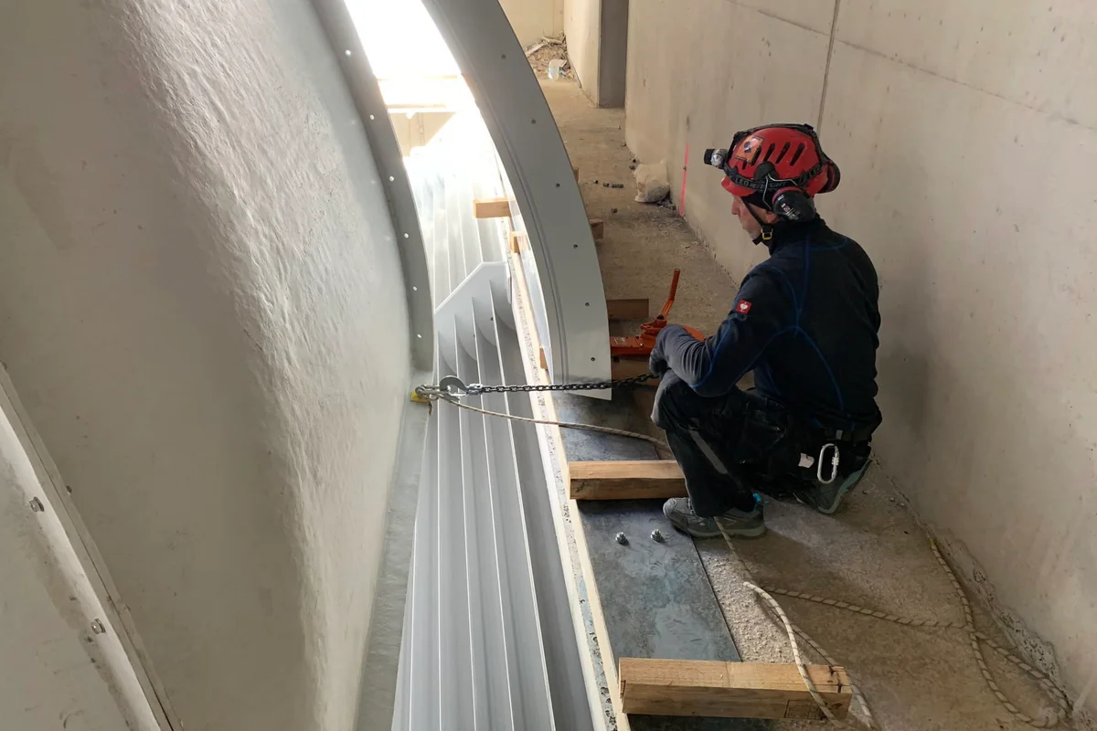 Een technicus positioneert een gebogen FRP-composietkanaalsectie met keerschoepen tijdens de montage van de Luxfly-windtunnel. Dit onderdeel vormt een hoek van het TT45 PRO recirculatiecircuit, ontworpen om de luchtstroom binnen de betonnen structuur om te leiden. Het installatieproces omvat een nauwkeurige uitlijning van het composietkanaalwerk met behulp van hijskettingen om in de bouwschil te passen.