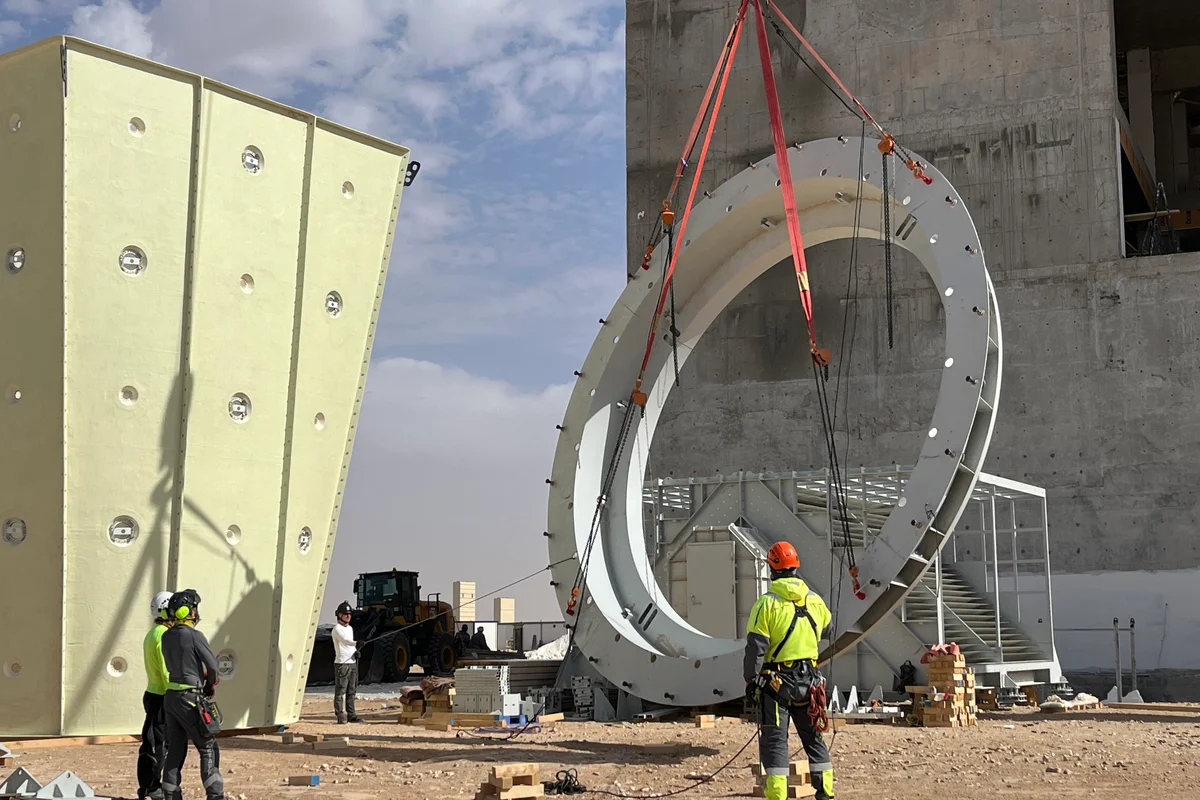 Wafanyakazi wa usakinishaji wanaongoza uinuaji wa kreini wa flange kubwa ya chuma ya kimuundo kwenye tovuti ya ujenzi wa handaki la upepo. Sehemu ya wima ya duct ya hewa ya kompositi ya FRP ya TunnelTech inasimama upande wa kushoto, ikiwa na muundo wa "sandwich" ulioundwa kwa ajili ya kunyonya kelele na ufanisi wa mtiririko. Mchakato wa kuunganisha unatumia vifaa vizito vya kuinua ili kuweka vipengele vya kipenyo kikubwa vya mzunguko wa aerodinamiki.