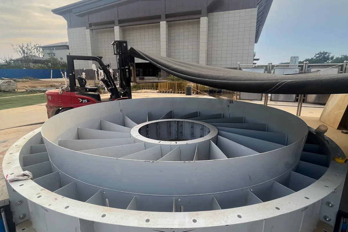 Duży, okrągły zespół łopatek stojana leży na ziemi podczas instalacji tunelu aerodynamicznego TT43 Smart w China Flight Town. Element ten posiada stałe, promieniowe łopatki kierujące rozmieszczone między wewnętrzną piastą a zewnętrzną obudową, zaprojektowane do zarządzania przep ływem powietrza w sekcji wentylatora osiowego. Logo TTE GMBH jest widoczne na zewnętrznej stronie stalowej obudowy. Sprzęt budowlany, w tym wózek widłowy i elektronarzędzia, otacza jednostkę, a w tle widać budynek obiektu.