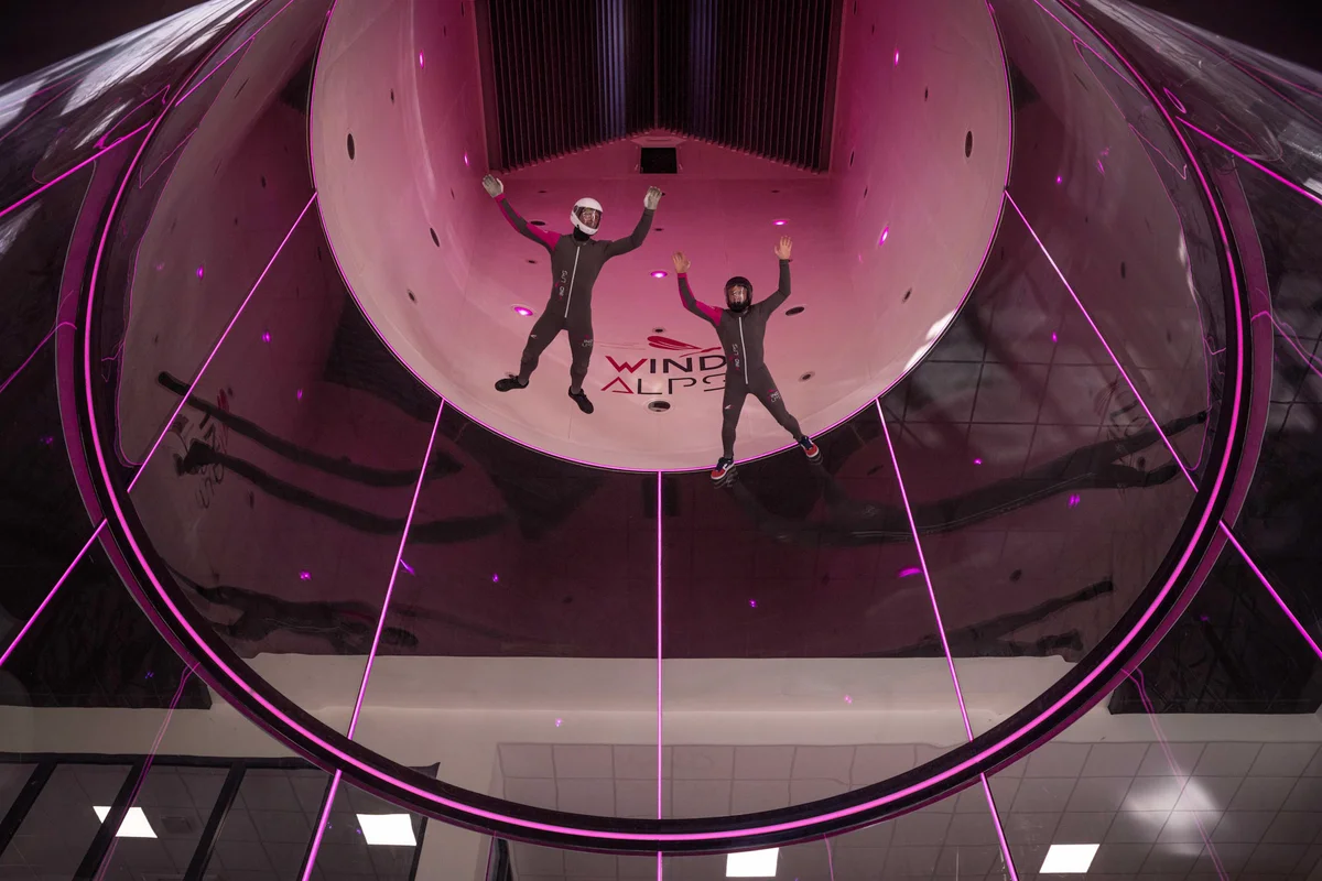 Two flyers execute belly flying maneuvers within the TT45 PRO flight chamber at the Wind Alps facility in France. The upward perspective showcases TunnelTech's engineering: a 4.5m diameter cylindrical section made of perfectly round, noise-absorbing multilayer glass. This frameless design provides distortion-free visibility, essential for safety and high-level professional coaching.