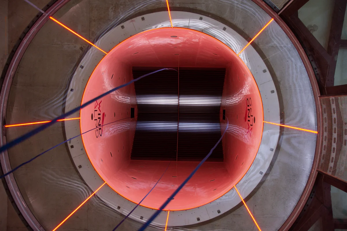 Une vue verticale regardant vers le haut à travers le circuit aérodynamique de l'installation Wind Alps en France pendant la phase d'installation. L'image montre la transition de la section circulaire au coin supérieur du conduit, où des aubes directrices horizontales sont positionnées pour rediriger le flux d'air. Des cordes de gréement bleues pendent au centre, utilisées pour l'accès sur cordes lors de l'assemblage de cette soufflerie TT45 PRO. Les murs intérieurs affichent la marque Wind Alps et des bandes d'éclairage LED radiales intégrées à la structure.