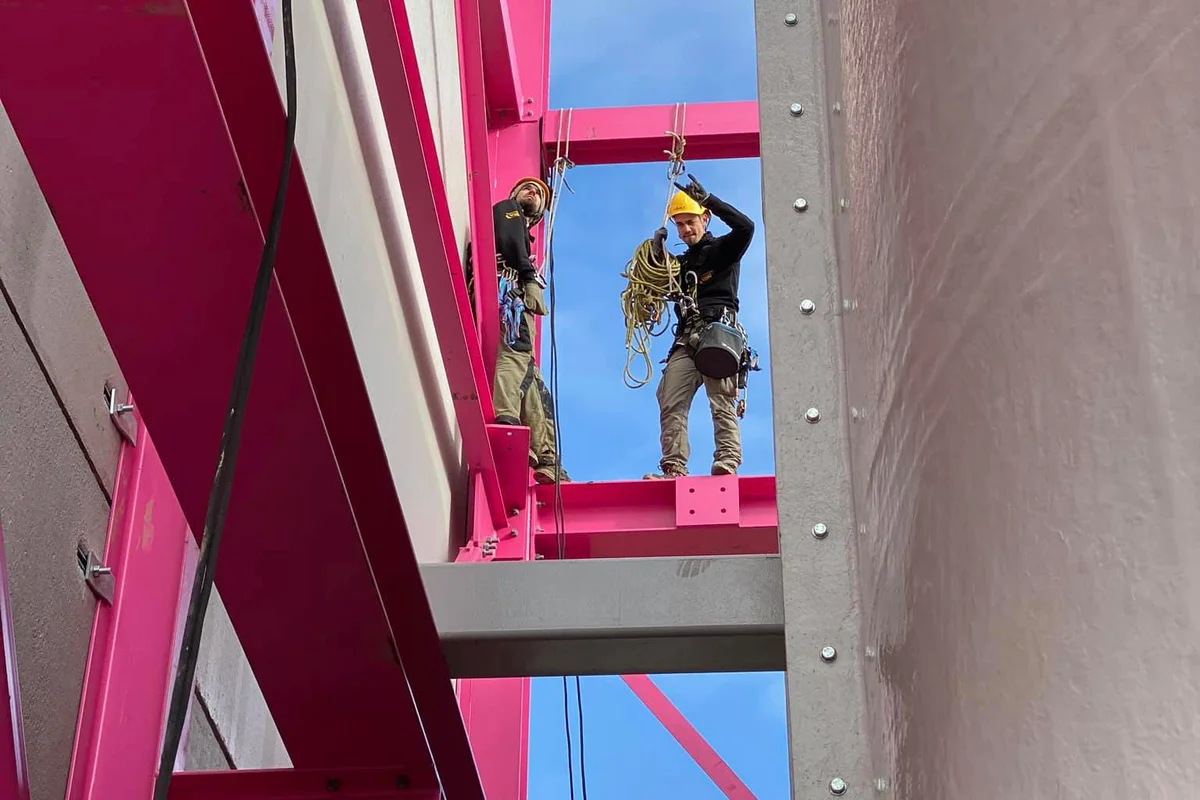 Kaks turvarakmete ja taglasevarustusega paigaldustehnikut seisavad Luksemburgis asuva Luxfly indoor skydiving rajatis ehituse ajal teraskarkassil. Töötajad teostavad kõrgustes taglastustöid, et joondada TunnelTech TT45 PRO tuuletunneli komponendid hoone iseloomuliku roosa kandekonstruktsiooniga. Paremal on näha vertikaalse tuuletunneli montaaži välispind terastalade kõrval.