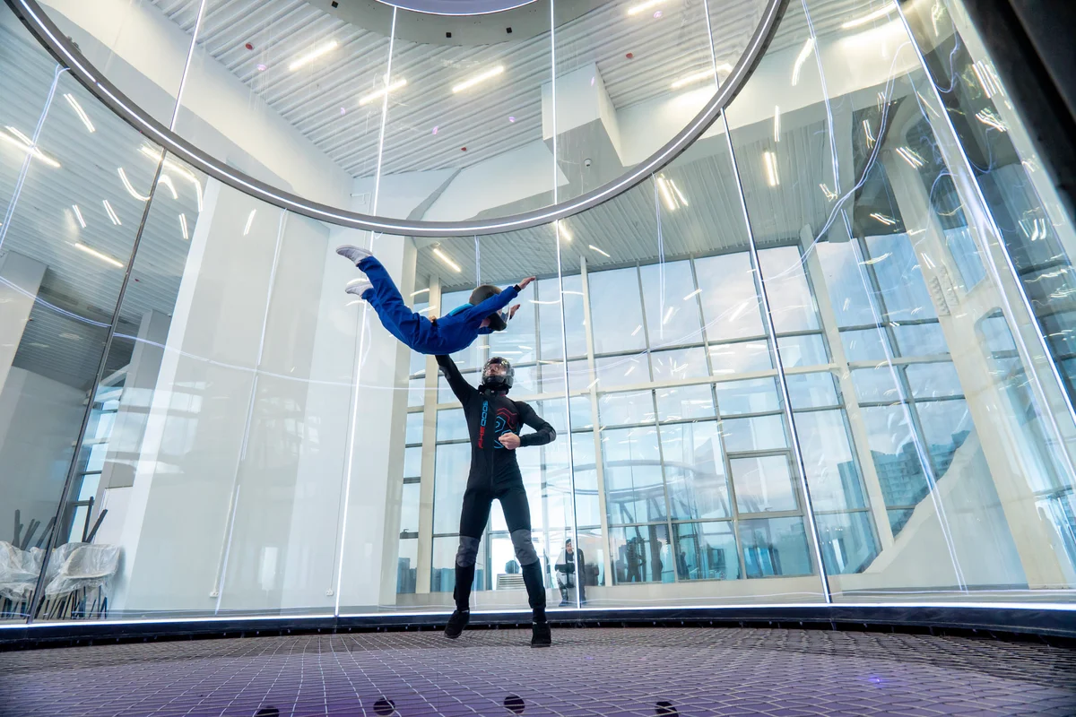 Een instructeur begeleidt een jonge vlieger tijdens een belly flying-sessie in de TT52 Pro windtunnel van de Kuzbas Arena. De vluchtkamer, met een diameter van 5,2 meter, is voorzien van gebogen meerlaags glas dat toeschouwers een perfect zicht biedt op de actie. Boven het stalen veiligheidsnet zorgt de instructeur via spotting voor de stabiliteit en correcte lichaamshouding van de leerling in de luchtstroom.