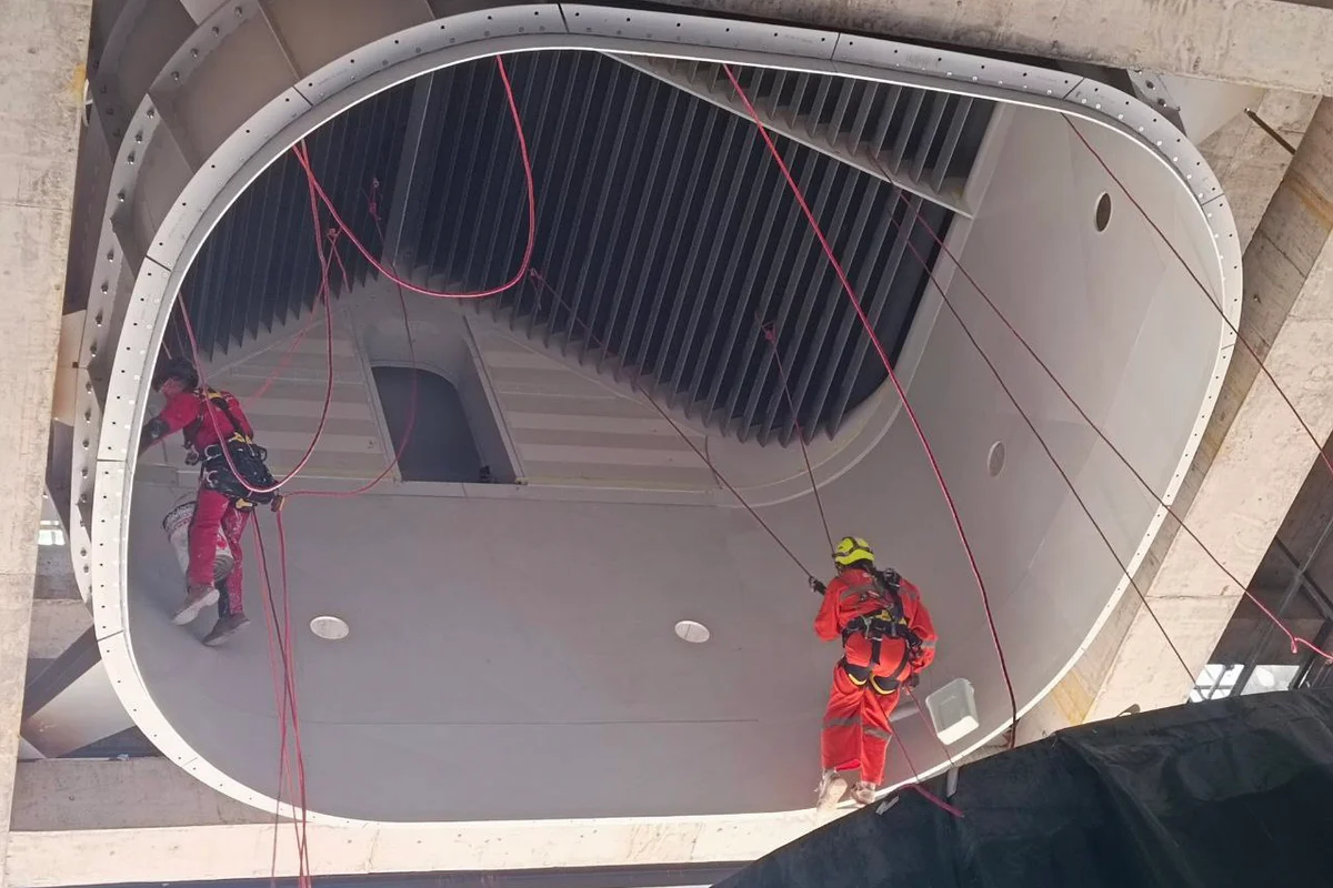 Teknikere utfører installasjon med tilkomstteknikk i den øvre delen av flykammeret for TT43 Smart-vindtunnelen ved China Flight Town. Bildet viser den hvite komposittstrukturen med sirkulære utskjæringer designet for installasjon av lyskilder. Over arbeiderne gir en rektangulær dør tilgang til BASE-hoppkammeret. Øverst i seksjonen er ledeskovler arrangert i en V-formet konfigurasjon for å splitte luftstrømmen til venstre og høyre returkanaler.