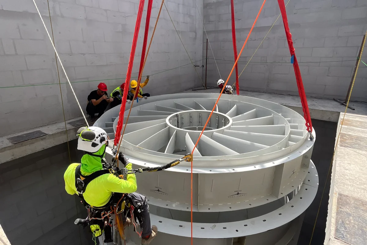 Un tecnico su fune guida l'installazione del gruppo delle pale dello statore del ventilatore assiale nell'alloggiamento in calcestruzzo. Questa operazione di sollevamento pesante fa parte della costruzione di una galleria del vento TunnelTech TT43 Smart presso il progetto China Flight Town. La sezione in acciaio contiene le pale fisse dello statore necessarie per raddrizzare il flusso d'aria all'interno del condotto di ritorno verticale. Il componente è sospeso da cinghie di sollevamento rosse mentre scende nella struttura in calcestruzzo dell'impianto.